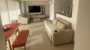 A modern living room with a large flat-screen TV, a beige sofa, a dining table with chairs, and a small wooden stool. - PONY PALACE Best Rooftop Pool in Cartagena! Beachfront, 3BR luxury condo balcony (Unknown)