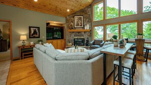 A cozy living room with a stone fireplace, wooden ceiling, and large windows offering a view of trees. - New! Epic Lake Views, Games, Hot Tub Comfort & A/C (Oakland)