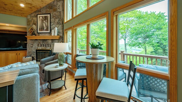 A cozy living room with a stone fireplace, comfortable seating, and large windows offering a view of the outdoors. - New! Epic Lake Views, Games, Hot Tub Comfort & A/C (Oakland)