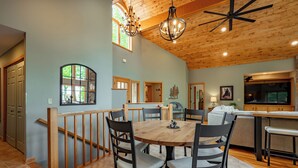 A spacious dining area with a wooden table and chairs, a chandelier, and a view of the kitchen. - New! Epic Lake Views, Games, Hot Tub Comfort & A/C (Oakland)