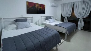 A room with two beds, a painting on the wall, an air conditioning unit, and a window with curtains.