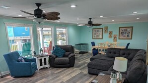 A living room with teal walls, a ceiling fan, and a dining area with a table and chairs. - 4 min walk Ocean View Sleeps 14.  Pet Ramp. Fenced Yard. King Bed 75" TV (Bolivar Peninsula)