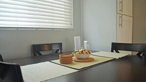 Une table à manger avec un saladier rempli de collations et un récipient blanc, entourée de chaises et d’une fenêtre avec des stores.