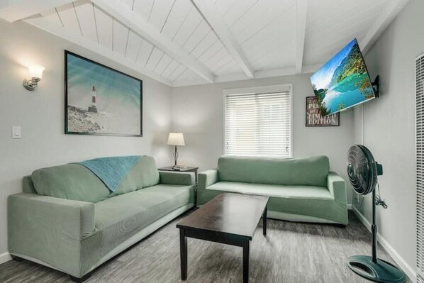 A living room with two green sofas, a wooden coffee table, a wall-mounted TV, and a framed picture of a lighthouse.