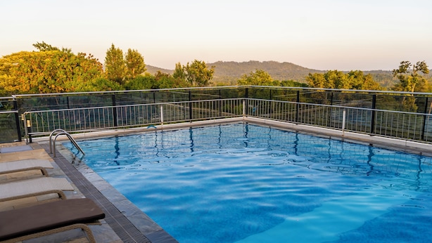 Una piscina rectangular con barandilla metálica, escalones de piedra para entrar y una vista de árboles y colinas al fondo.
