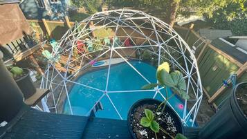 Une piscine dotée d’une structure en forme de dôme géodésique, entourée de plantes en pot et de mobilier d’extérieur.