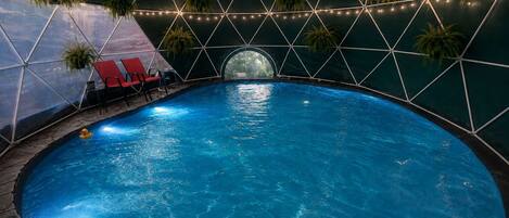 An indoor swimming pool with a geometric ceiling structure, surrounded by lounge chairs and potted plants.