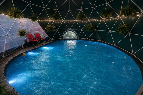 An indoor swimming pool with a geometric ceiling structure, surrounded by lounge chairs and potted plants.