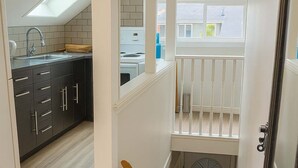 A modern kitchen with dark cabinets and a white countertop, a wooden staircase leading to a second floor, and a view of the outside through a window. - Quaint Attic Suite w/ Heated Pool (Year-Round) (Victoria)