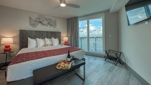 Une chambre d’hôtel avec un grand lit, une table de chevet avec une bouteille et un verre, un plateau de collations, un ventilateur de plafond et une vue sur le ciel et l’eau par une grande fenêtre.