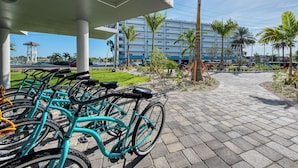 Une rangée de vélos turquoise est garée sous un abri, d’où l’on peut admirer un immeuble à plusieurs étages et des palmiers en arrière-plan.
