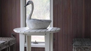 Una habitación rústica con una mesa blanca que presenta una escultura de cisne, una ventana y dos sillas de mimbre.