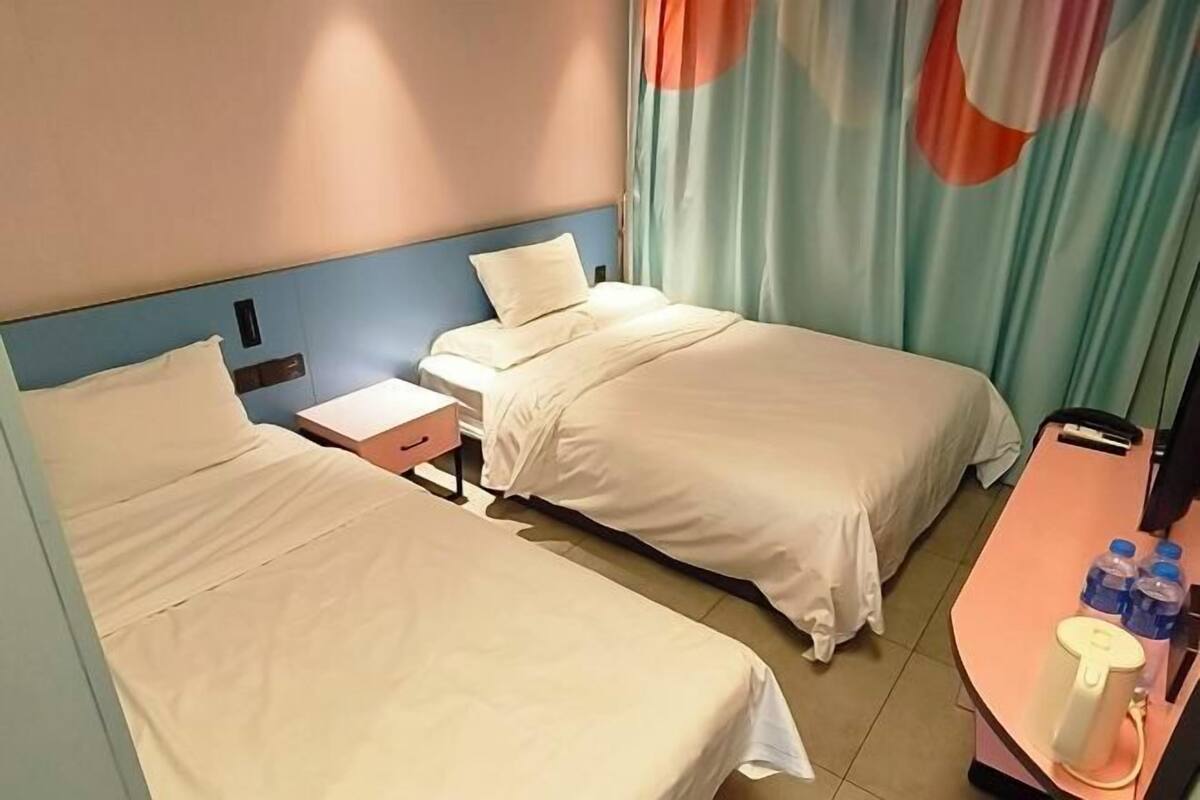 A hotel room with two beds, a nightstand, a small table with a lamp, and a window with curtains.