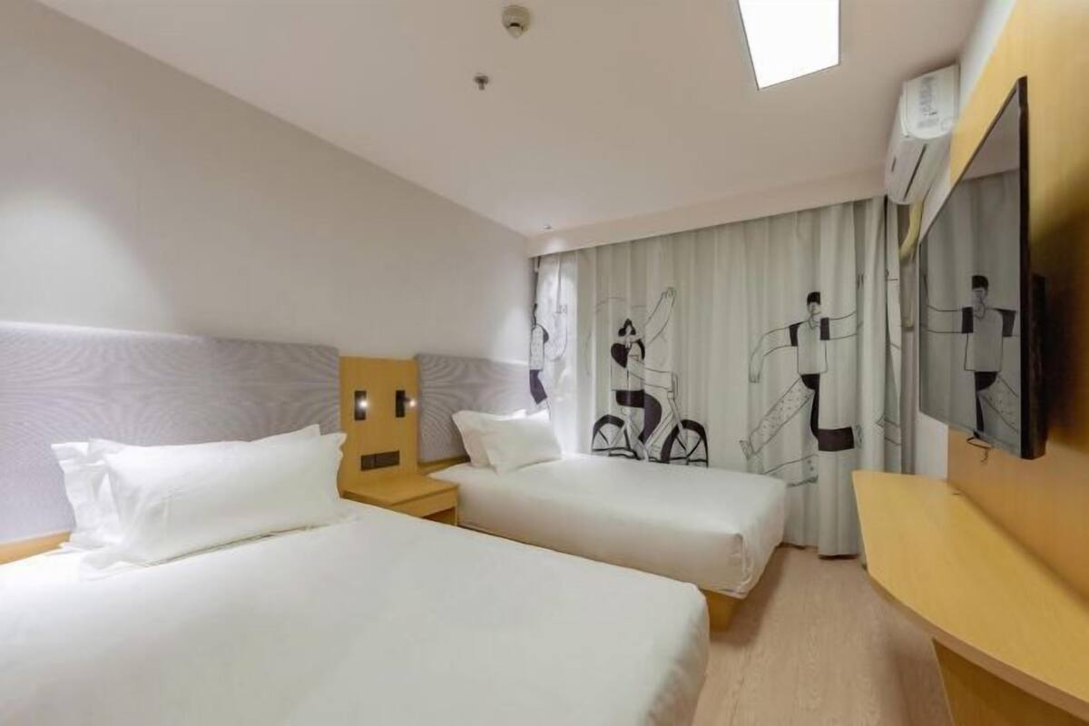 A hotel room with two beds, a TV, a desk, and a window with curtains.