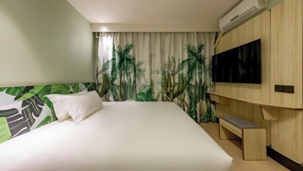 A hotel room with a bed, a bench, a TV, and an air conditioning unit.