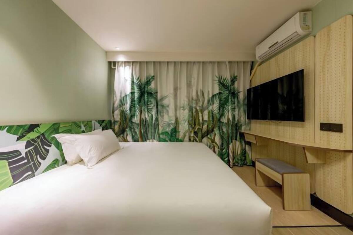 A hotel room with a bed, a bench, a TV, and an air conditioning unit.