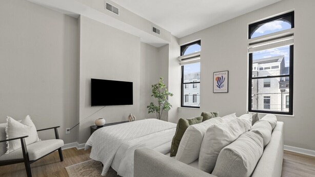A modern bedroom with a large bed, a sofa, a TV, and two windows offering city views.