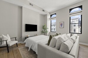 A modern bedroom with a large bed, a sofa, a TV, and two windows offering city views. - The Grand Flat | 1800 sq ft | Free Parking | OTR (Cincinnati)