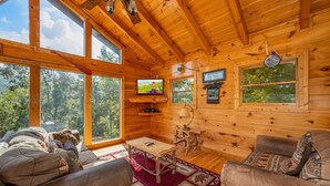 L’intérieur chaleureux d’un chalet avec des murs en bois, de grandes fenêtres, un téléviseur, une table et des sièges confortables.