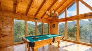 Une table de billard en bois dans une pièce dotée de grandes fenêtres et d’un lustre.
