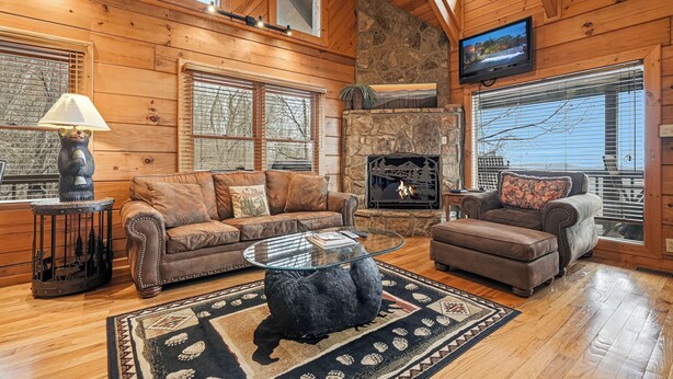 A cozy living room with wooden walls, a stone fireplace, a television, and a large window.