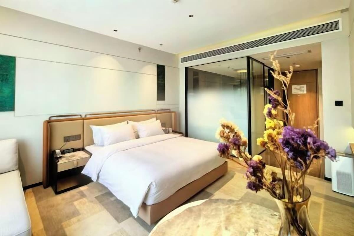 A modern hotel room with a large bed, bedside tables, a nightstand, a vase with flowers, and a sliding glass door.