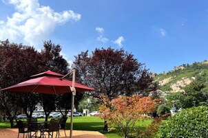 An outdoor seating area with a red umbrella, tables, and chairs, surrounded by trees and a hilly landscape.