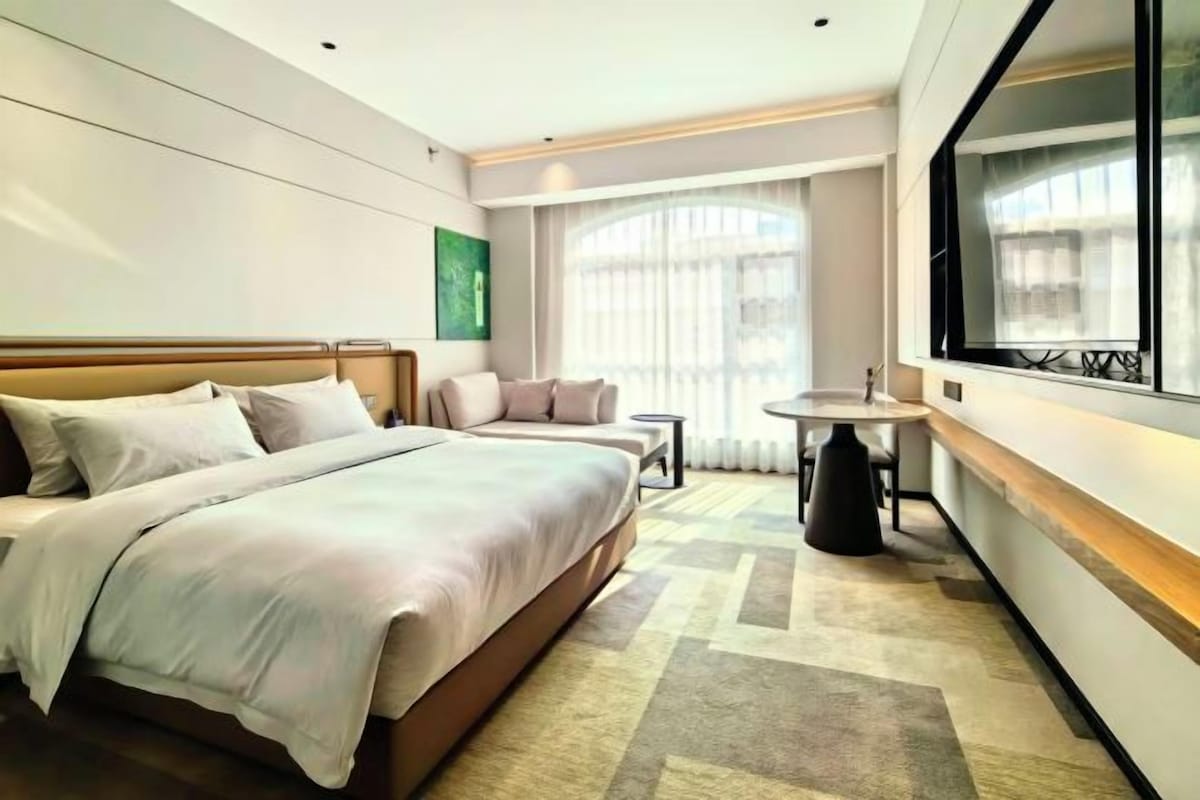 A modern hotel room with a large bed, a sofa, a small table, and a large window.