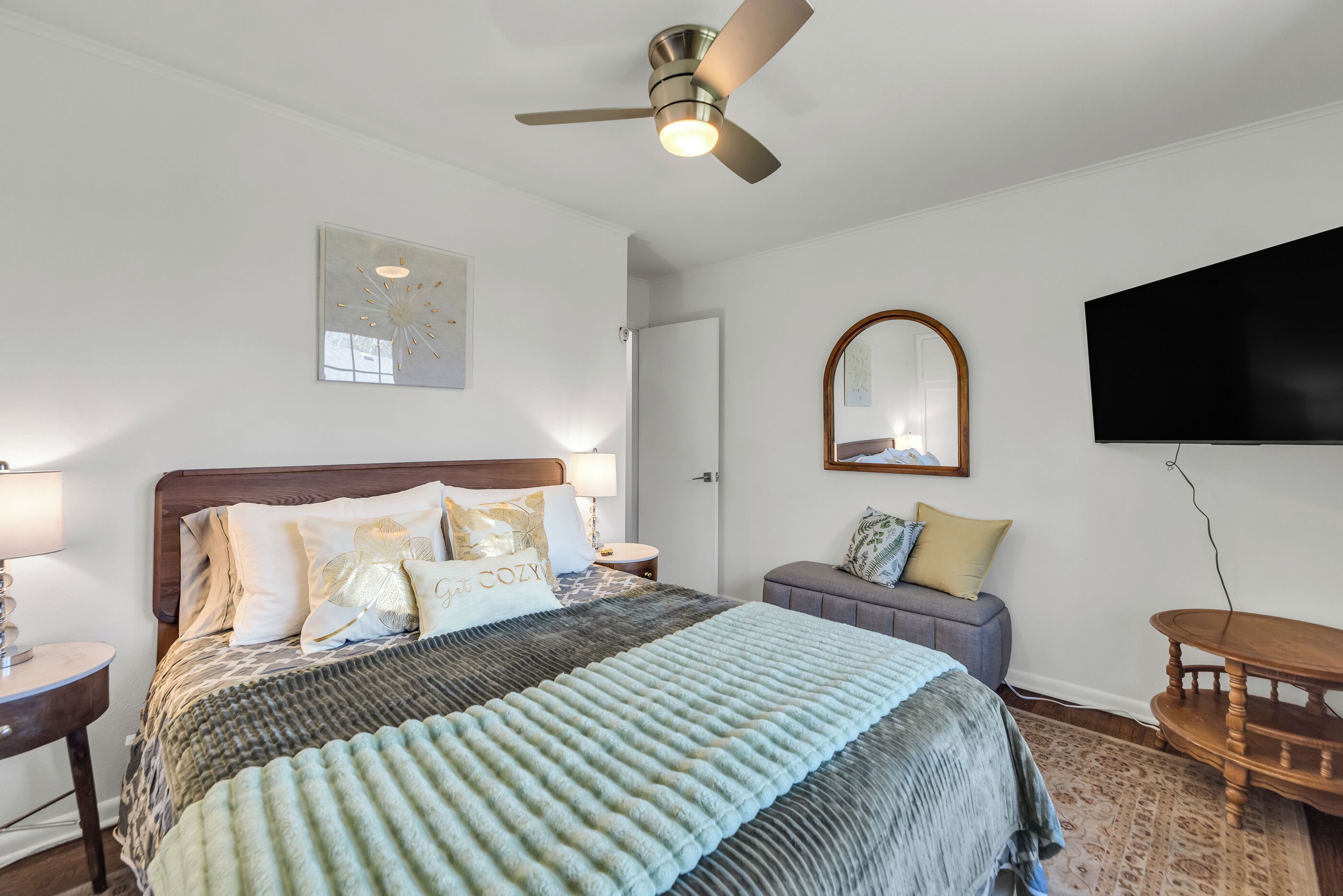 A bedroom with a bed, a ceiling fan, a TV, a mirror, and a small table.