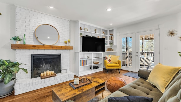 Una sala de estar con chimenea de ladrillo blanco, televisor de pantalla plana, un sillón amarillo y una mesa de centro de madera.
