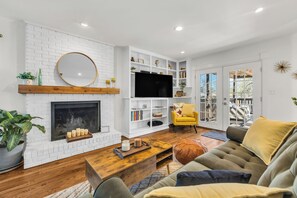 Un salon avec une cheminée en briques blanches, un téléviseur à écran plat, un fauteuil jaune et une table basse en bois.
