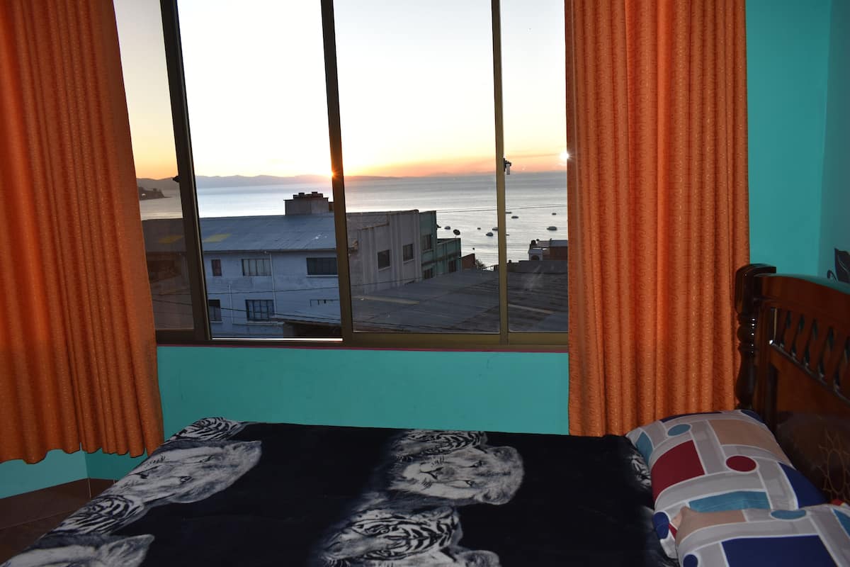 A bedroom with a bed, a window with curtains, and a view of the sea and buildings.