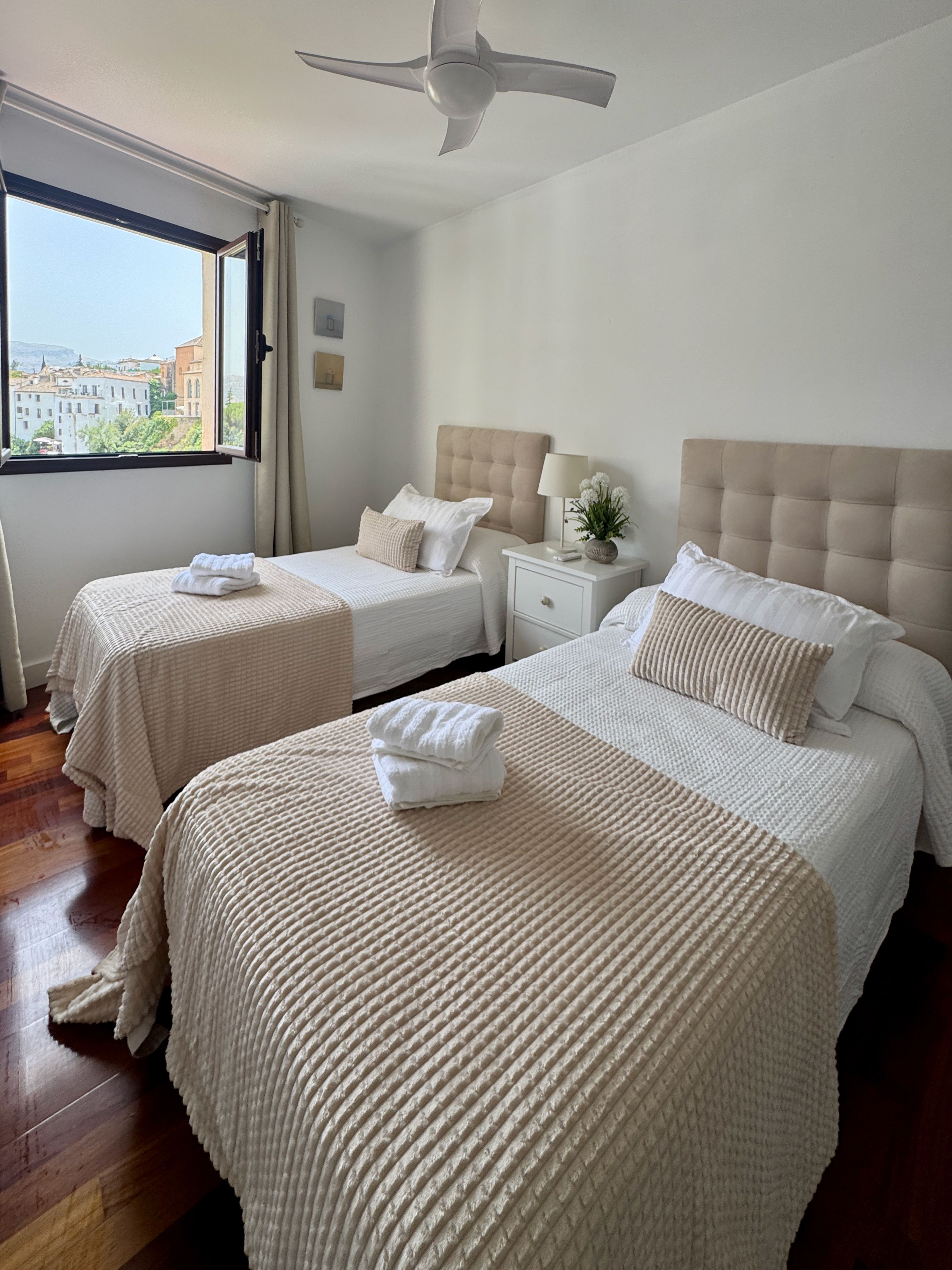 A hotel room with two beds, a ceiling fan, a nightstand with a lamp, and a window with a city view.
