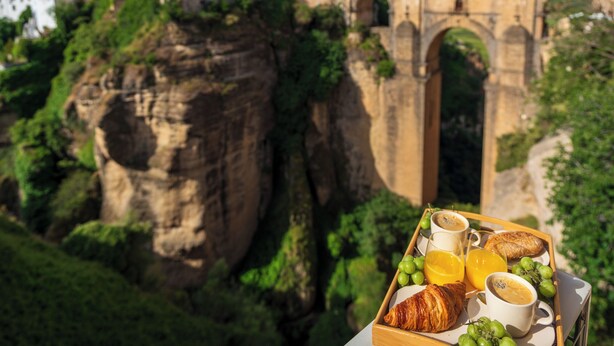 A breakfast tray with croissants, grapes, orange juice, and coffee, set against a backdrop of a stone bridge and buildings. - Las Vistas Residence - Best Views in Ronda (Ronda)