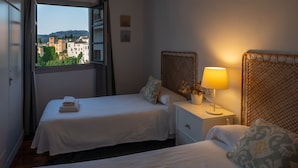 Habitación de hotel con dos camas, una ventana con vistas a edificios, una mesita de noche con lámpara y una planta pequeña en maceta.