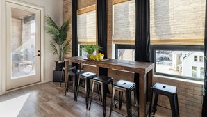 Een moderne keuken met een houten bar en krukken, grote ramen met zonwering en een potplant.