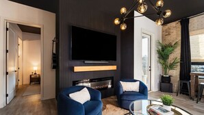 Een moderne woonkamer met twee blauwe fauteuils, een flatscreen televisie, een open haard en een glazen salontafel met boeken en een plant.