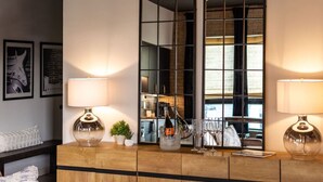 Een moderne woonkamer met een houten console, twee vloerlampen, een kleine potplant en een grote spiegel die een bakstenen muur en raam weerspiegelt.