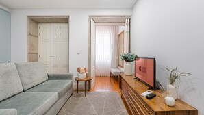 A modern living room with a grey sofa, a small round table with a vase of flowers, a wooden console table with a TV and remote, and a hallway leading to a door. - GuestReady - Lovely heaven in Porto (Porto)