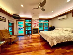A bedroom with a large bed, a ceiling fan, a fireplace, and a view through the windows.