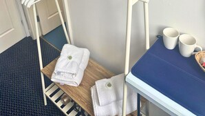 Habitación de hotel con televisor montado en la pared, un perchero blanco con ganchos, un pequeño estante de madera con toallas dobladas y una mesa con dos tazas blancas y una cesta.