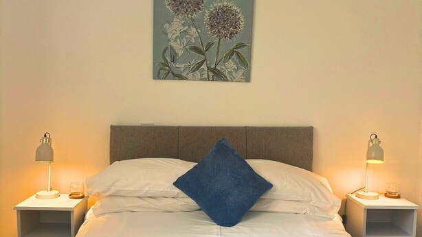 Una cama bien tendida con sábanas blancas y una manta azul, con dos mesitas de noche con lámparas a cada lado, frente a una pared lisa con una obra de arte floral sobre la cama.