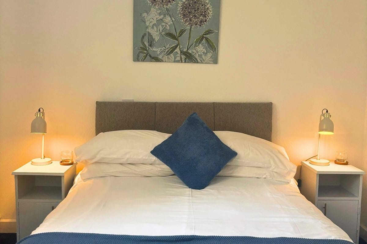 A neatly made bed with white linens and a blue throw blanket, flanked by two bedside tables with lamps, against a plain wall with a floral artwork above the bed.
