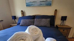 A neatly made bed with two rolled towels, a dark blue bedspread, and a painting of a lighthouse on the wall. - Mulberry House - Modern Family Retreat (Sligo)