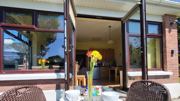 Un área de comedor al aire libre con sillas de mimbre, una mesa de vidrio, un taburete de madera y un jarrón con flores.