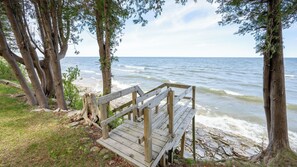 A wooden bench by a rocky shoreline with trees and a view of the ocean. - Lakefront Escape | Fire Pit, Pets & Scenic Views (Sturgeon Bay)