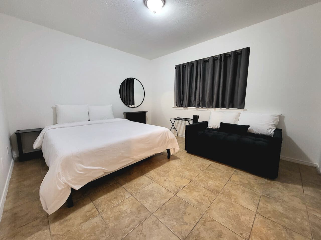 A bedroom with a bed, a black sofa, a round mirror, a window with curtains, and a tiled floor.