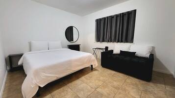 A bedroom with a bed, a black sofa, a round mirror, a window with curtains, and a tiled floor.