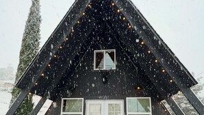 Una cabana negra amb un teulat triangular, una porta blanca i una finestra petita. Està caient neu i hi ha llums de garlandes al teulat.