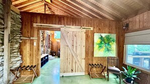 Ein rustikales Interieur mit Holzwänden, einer Steinsäule links, einer großen Holztür, einer Bank, einem Stuhl und einem Blumentopf.